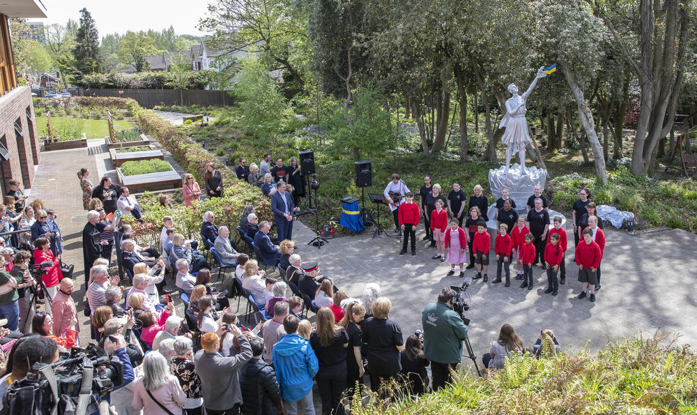 A crowd of people in the garden at Strawberry Field watch a choir in front of a new monument