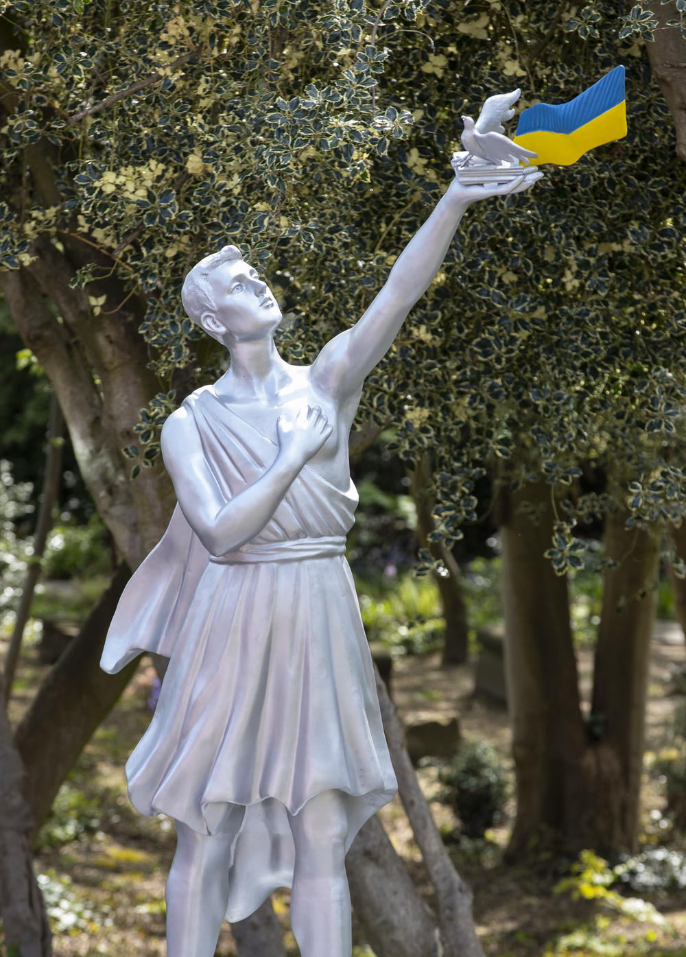 Aluminium statue of a man holding a flag, dove and book aloft in a garden.