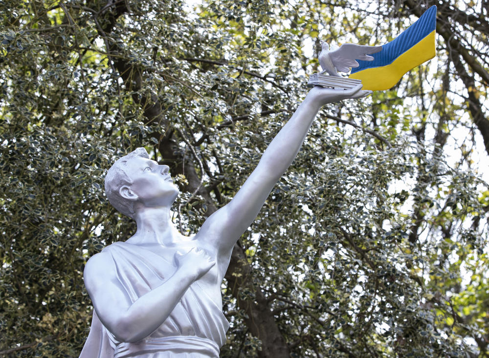 Close up of the upper part of a statue which shows a man holding a dove book and Ukrainian flag aloft with trees behind