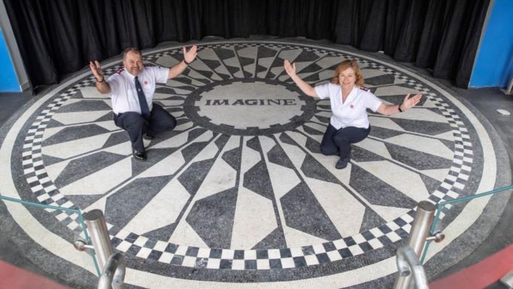 Two people in Salvation Army uniforms stand on a mosaic which features the word "Imagine" in tiles at Strawberry Field Liverpool UK